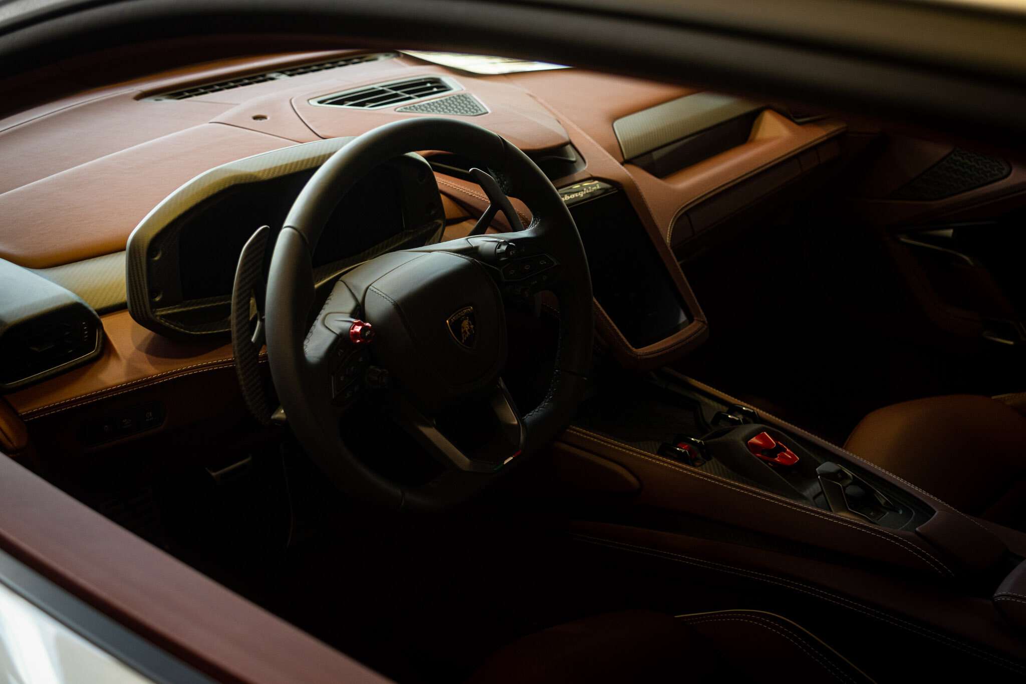 Close up of a Lamborghini's tan leather dashboard, carbon fiber trim, and steering wheel with red paddle shifters