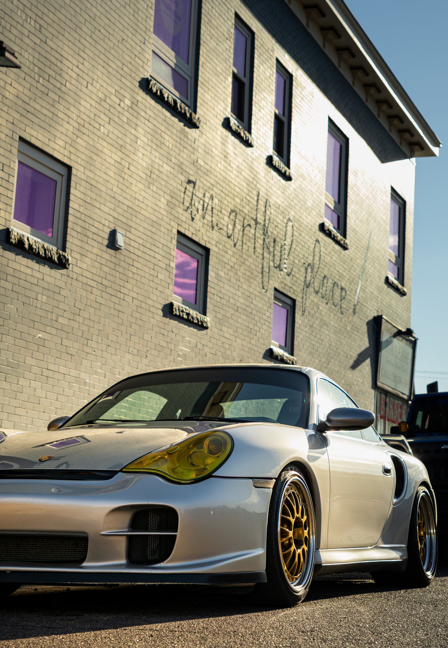Silver widebody Porsche 911 with gold wheels parked on the street in front of a building with a hand-painted mural