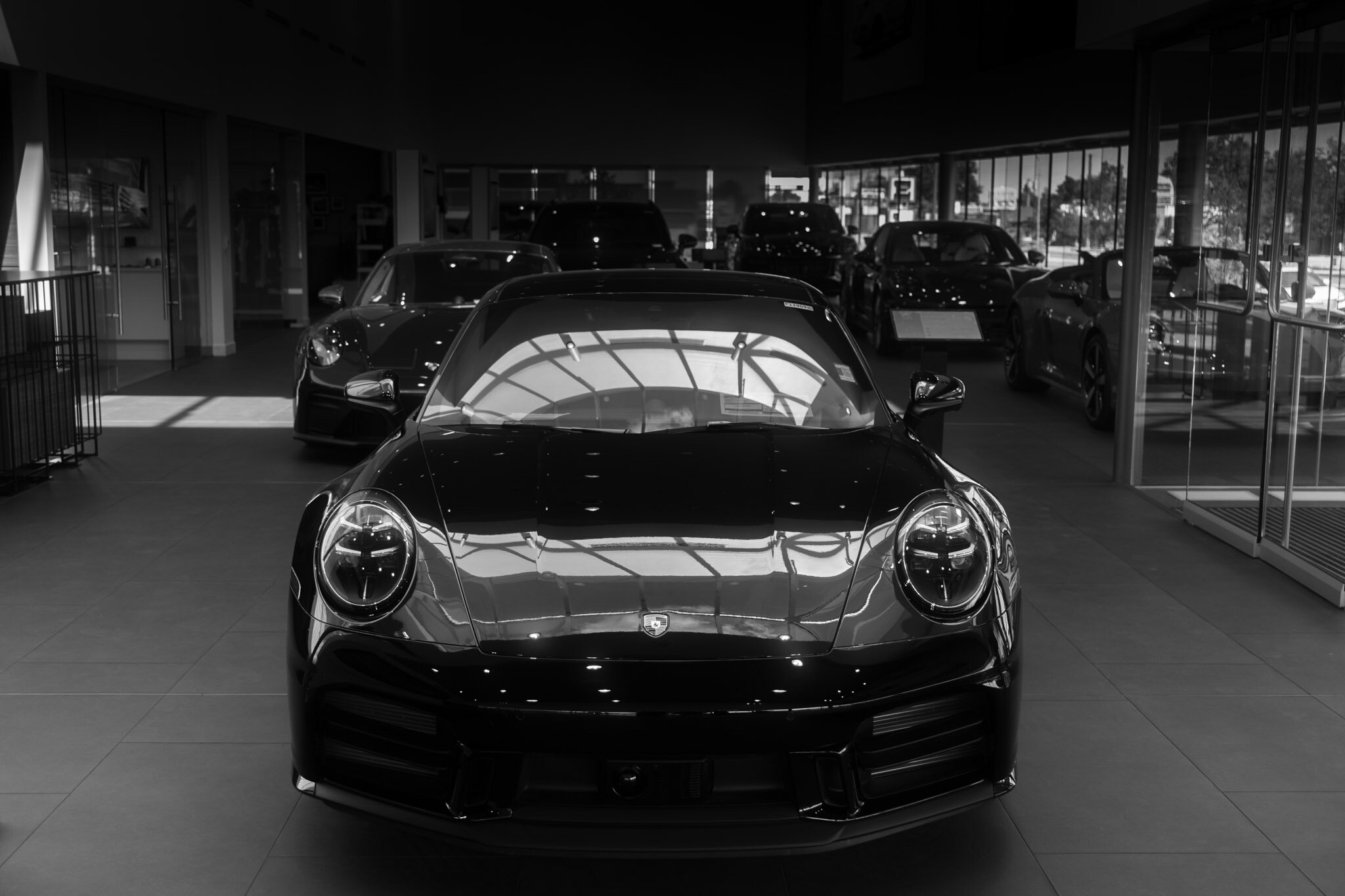 Black and white photo of a Porsche 911 head on inside a showroom, with several more cars reflected and lined up behind it